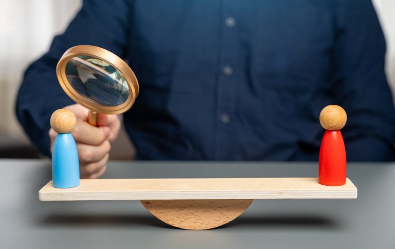 Man holding magnifying glass over two wooden people on a see saw depicting the comparison between Managed IT Services vs. Break-Fix IT